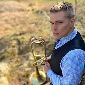 Young white man in blue checkered shirt and navy vest looking left to camera, holding a trombone