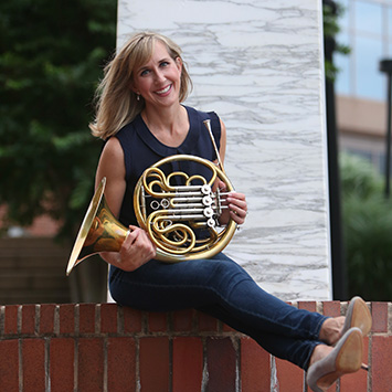 white woman in dark vest and dark jeans sitting on brick wall holding a French horn