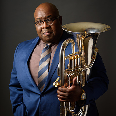 Headshot of man in blue suit against gray background, posing for portrait with euphonium in right hand.