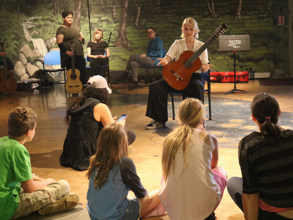 White female with blonde hair playing classical guitar instructs group of young students.