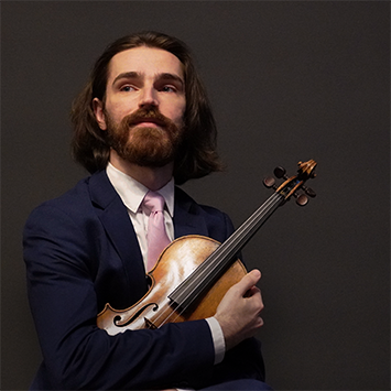 Portrait of Nathan Lowman in suit and pink tie against a slate gray background. Nathan is cradling his violin in his right hand and looking slightly to his right off camera.
