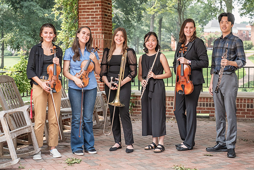 Group photo of 2023's concerto competition winners, all posing with their instruments. They are posed underneath Guilford College's brick gazebo with green summer foliage in the background.